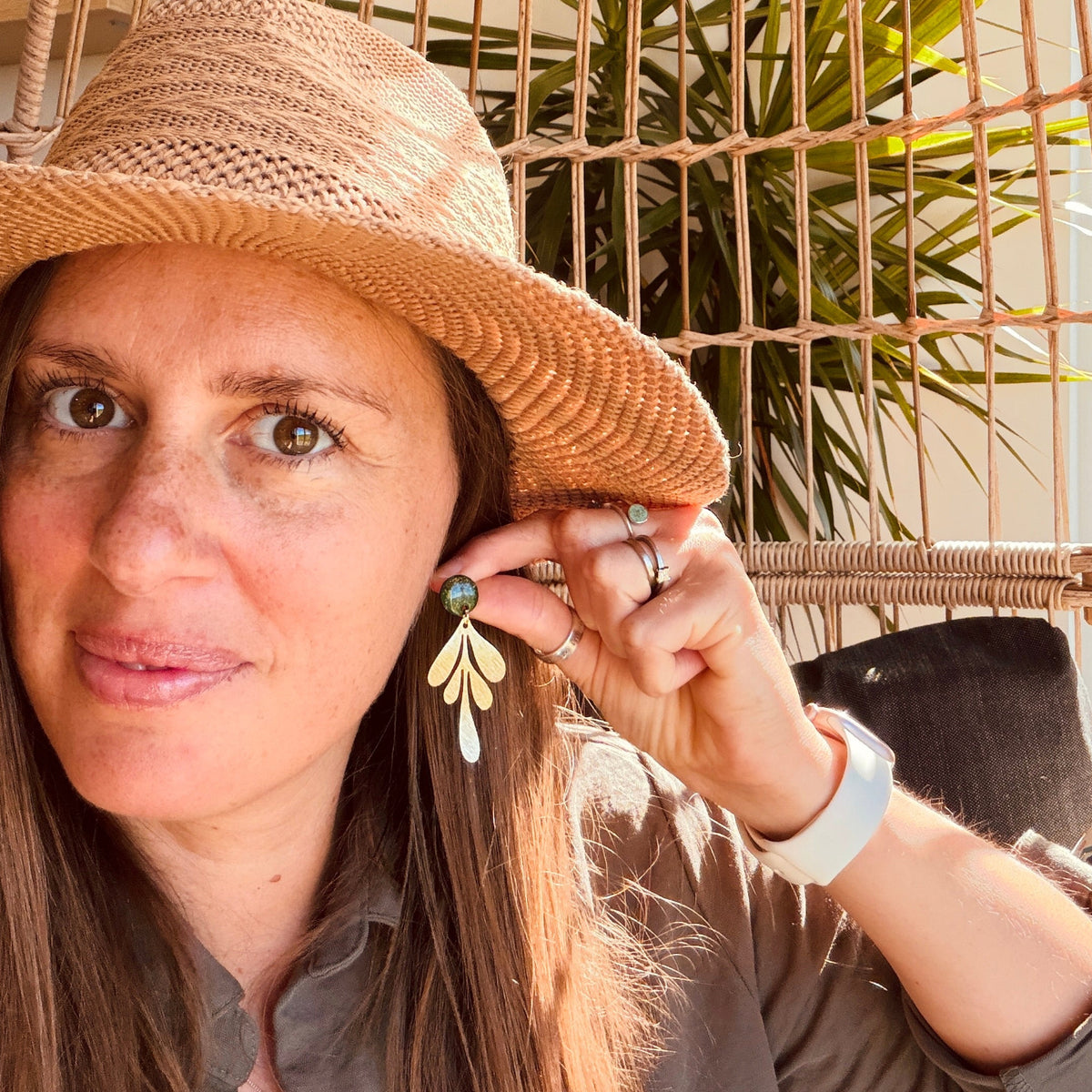 A woman wearing a straw hat and a gray shirt holds up a dangling leaf-shaped earring with a green stone and white leaf details, set against a wicker chair and green plants.