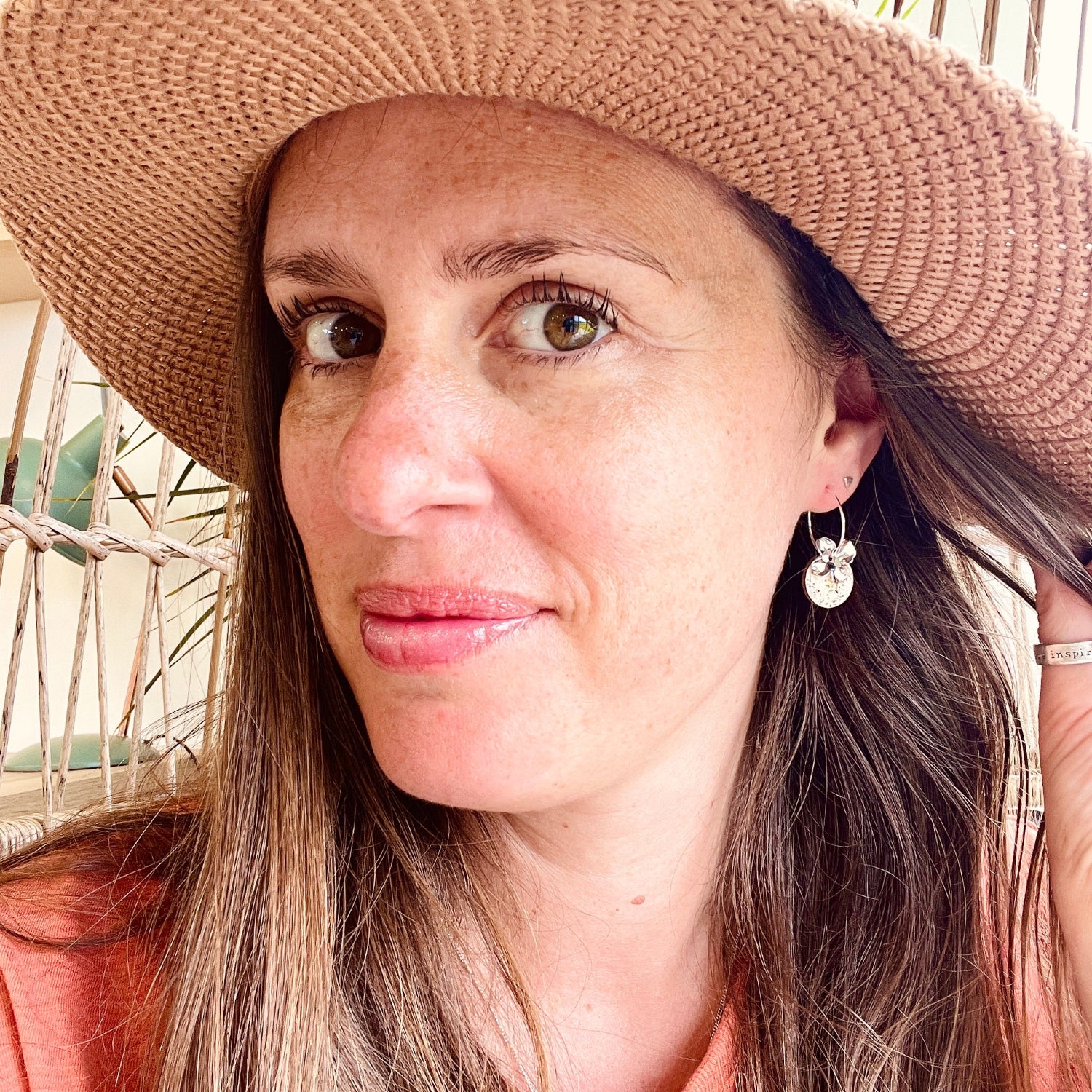 A woman wearing a wide-brimmed woven straw hat in a warm beige/tan color, photographed in a close-up portrait. The background shows a light bamboo or rattan wall.