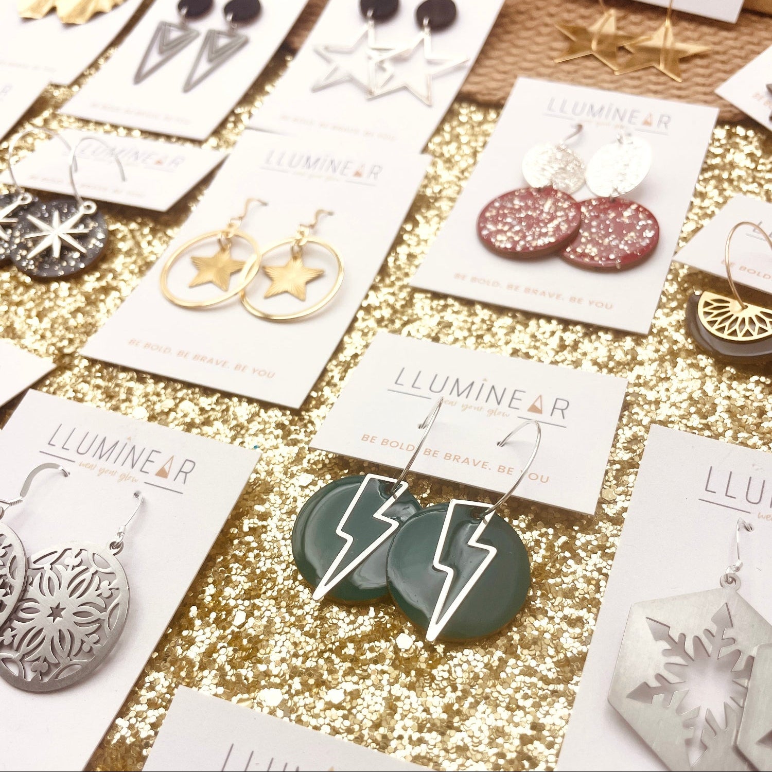 Various earrings on display with 'LLUMINEAR' branding on a glittery surface.