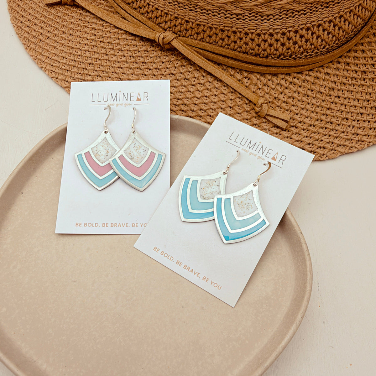 Two pairs of geometric drop earrings on white display cards, styled on a beige tray with a straw hat in the background. The earrings feature layered fan shapes in turquoise blue, dusty pink, and white.