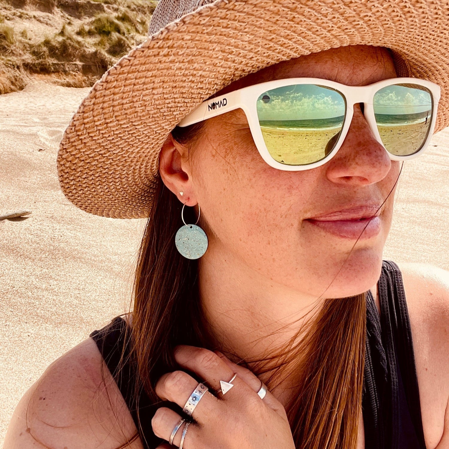 Woman wearing a straw hat and beach sand earrings with sunglasses sitting on a beach