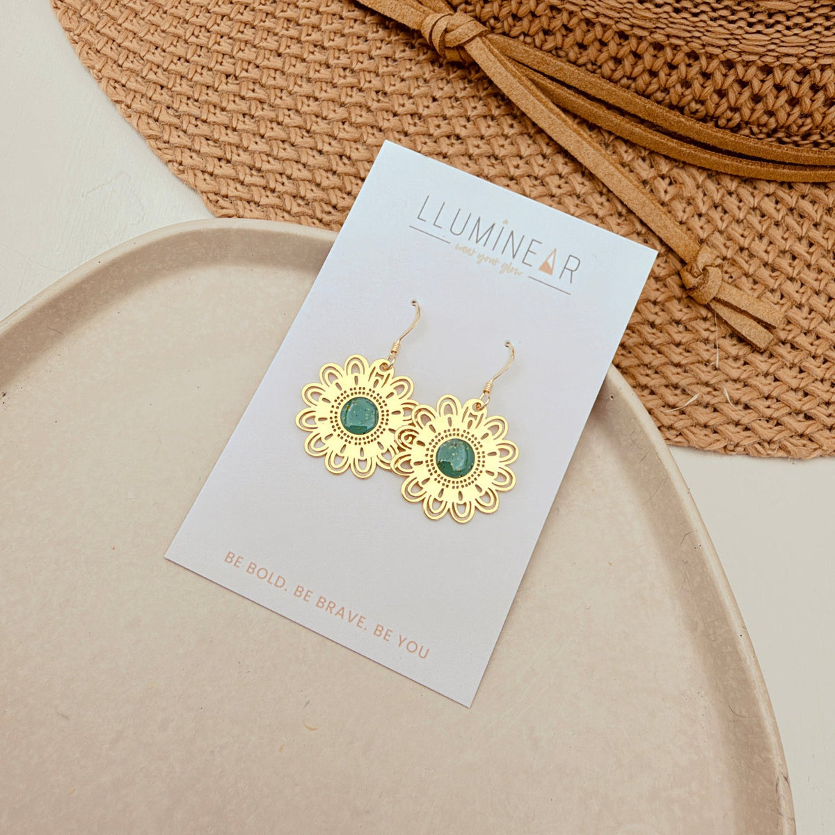 A pair of gold flower-shaped dangle earrings with green oval centers, displayed on a white card labeled 'ILLUMINEAR'. The card is placed on a beige tray, with a woven straw hat in the background.