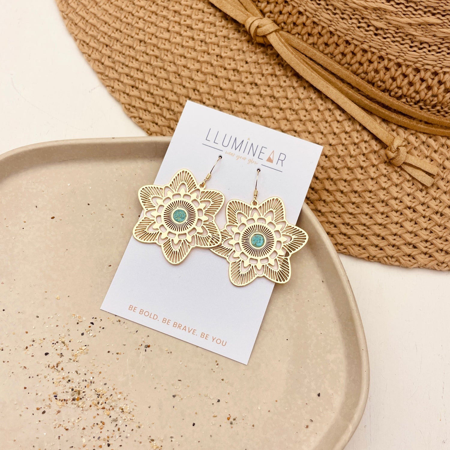 A pair of gold-tone flower-shaped dangle earrings with a small turquoise center, displayed on a white card labeled 'LLUMINE' and placed on a beige tray next to a woven straw hat.