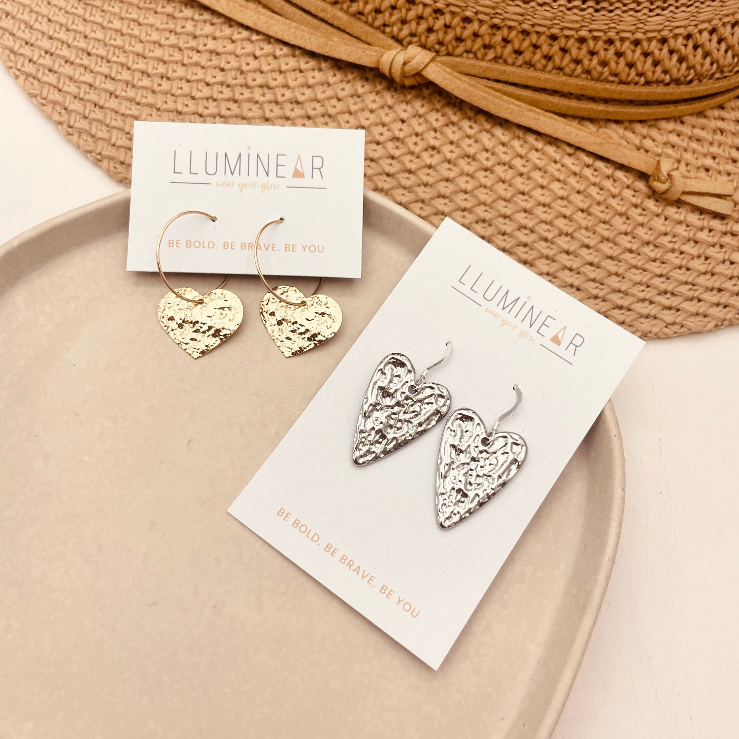 Heart-shaped earrings on a card with 'Lumineer' branding, placed on a beige plate with a woven mat in the background.