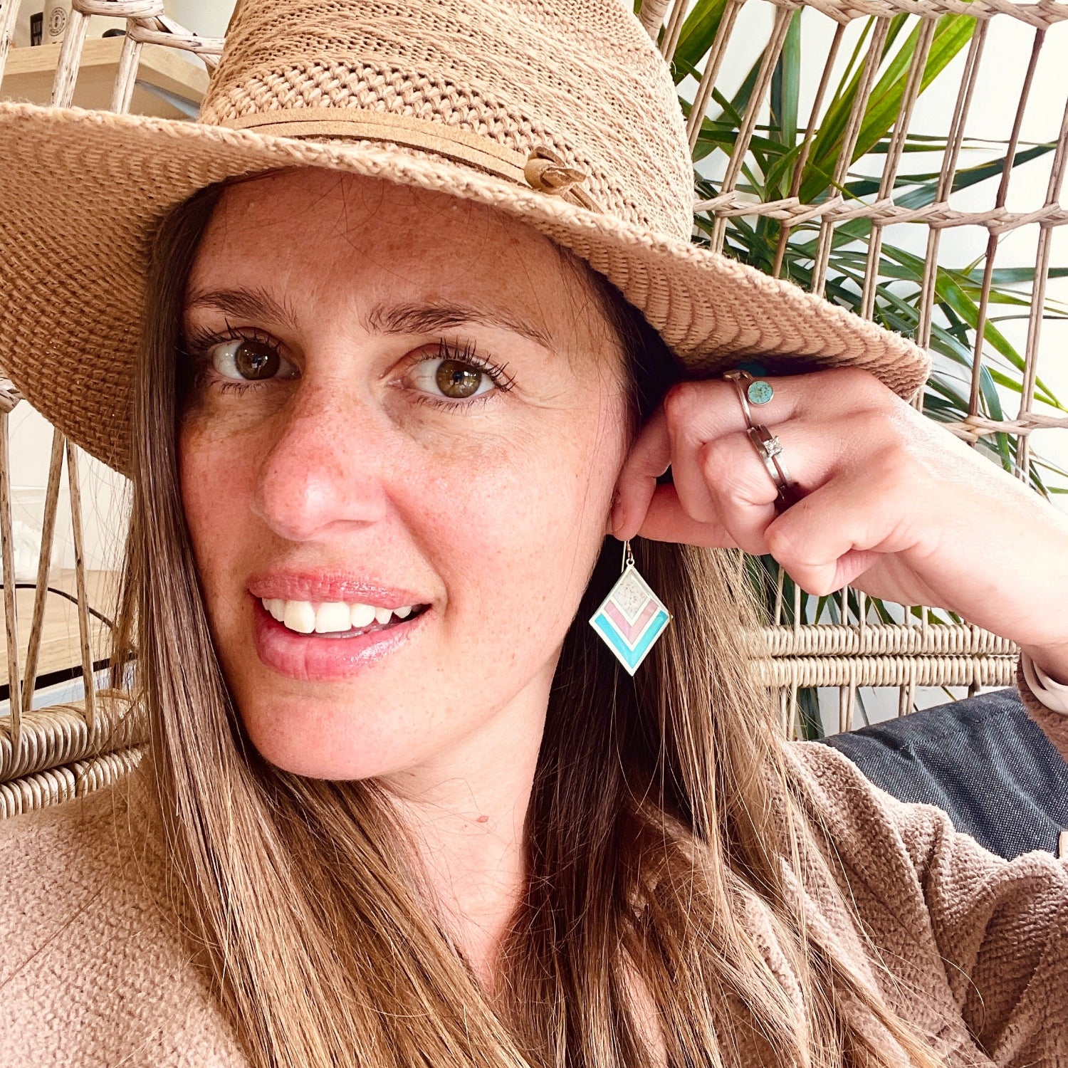 A woman wearing a wide-brimmed straw hat and a beige knit top, smiling while touching the brim of her hat. She is wearing a light blue and white diamond-shaped dangle earring.