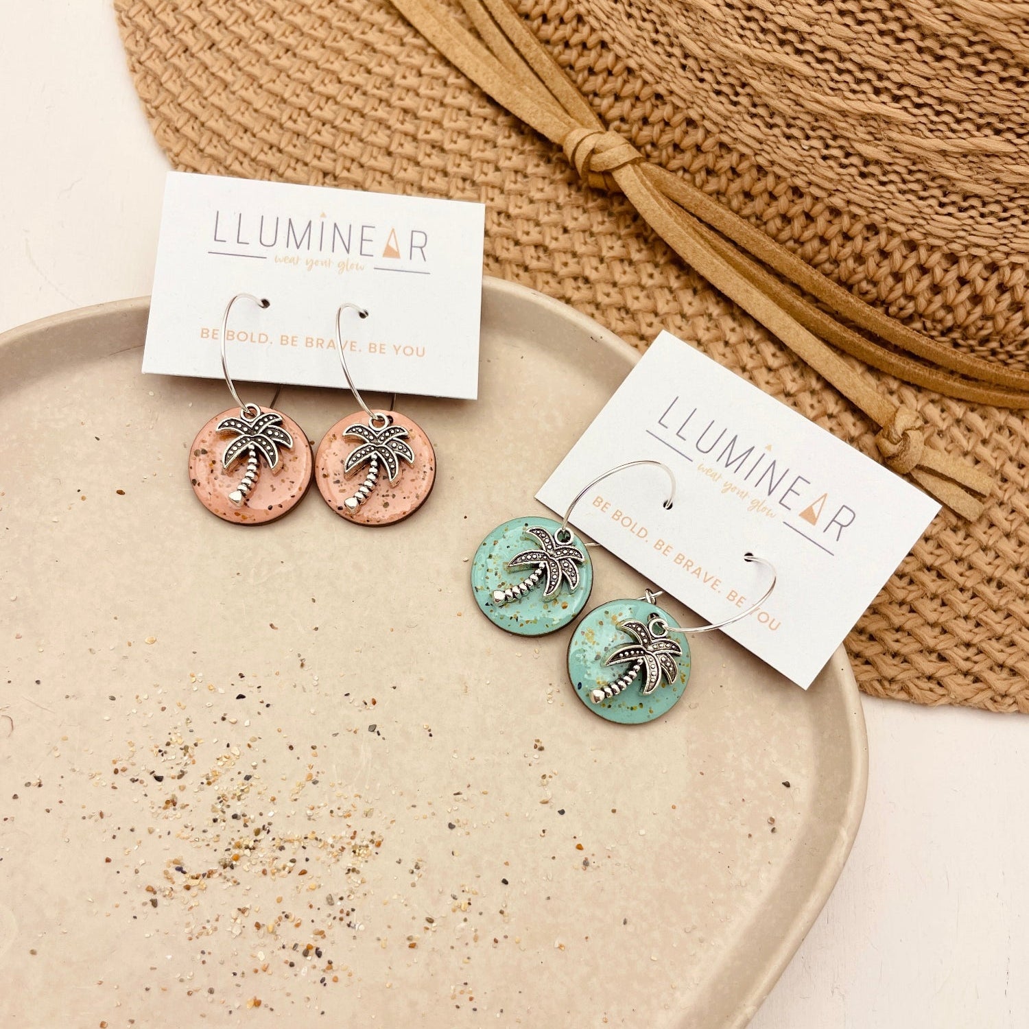 Two pairs of LlumineR palm tree dangle earrings displayed on white cards atop a speckled beige tray, with a woven straw hat in the background.