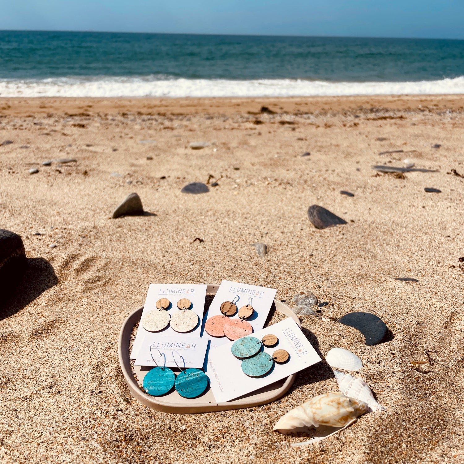 Set of earrings on a display card on a sandy beach with ocean in the background