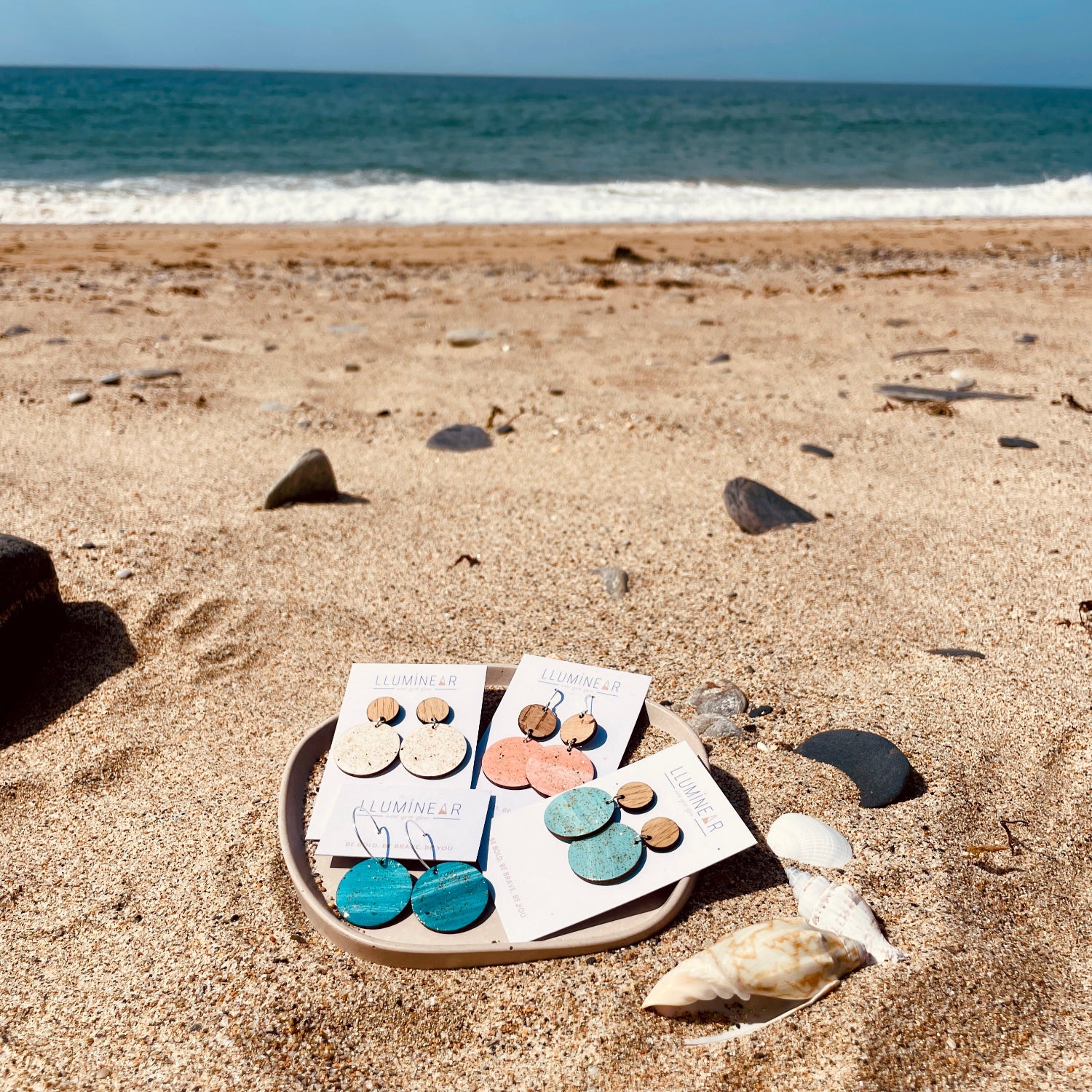 Set of earrings on a display card on a sandy beach with ocean in the background