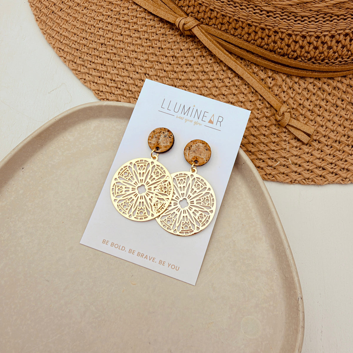 A pair of gold-toned dangle earrings with brown wooden beads and intricate filigree cutouts, displayed on a white brand card labeled 'LLUMINEAR'. The card is styled on a beige ceramic dish with a woven straw hat in the background.