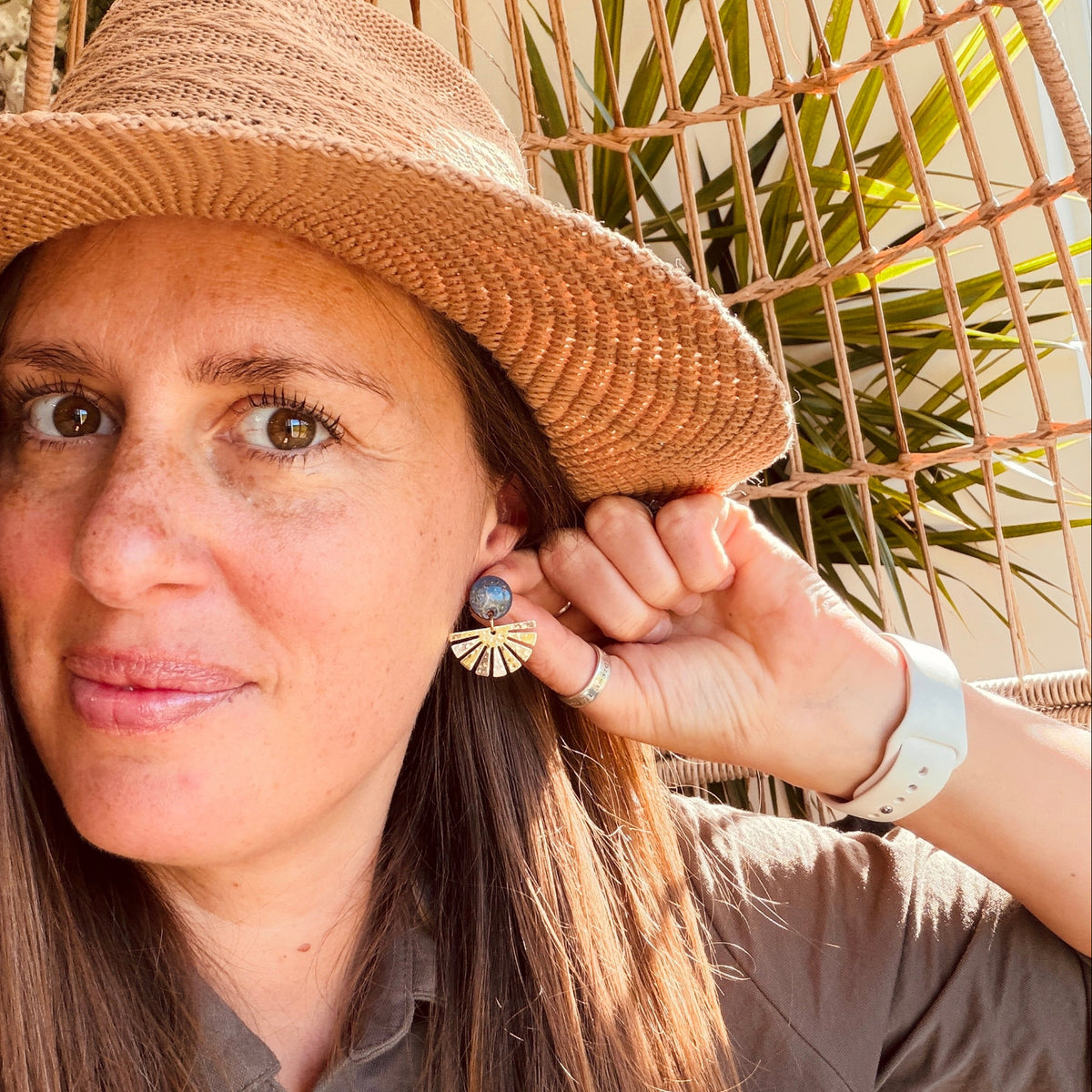 A woman wearing a straw hat and brown shirt models a pair of bohemian-style earrings with blue and gold details, set against a warm, sunlit wicker and plant background.