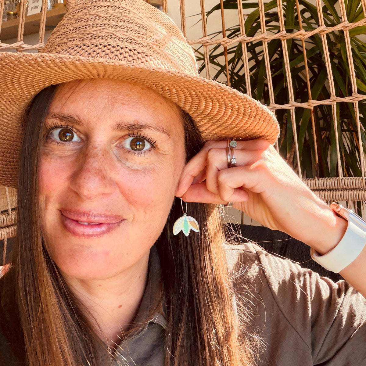 A woman wearing a straw hat and a brown shirt, holding her ear with her hand to show a small dangling earring. She is under a large woven wicker umbrella in a sunlit indoor setting with green plants in the background.