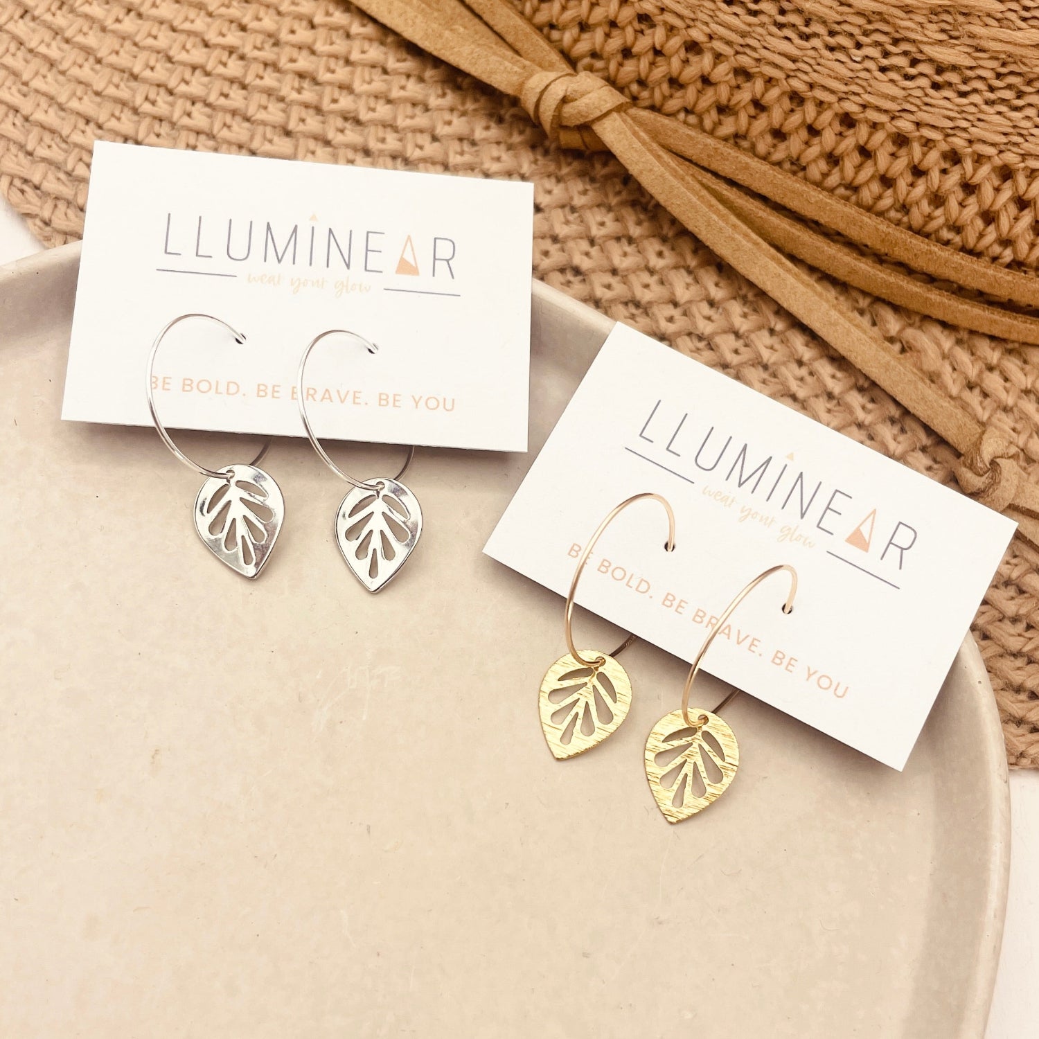 Gold leaf earrings on Lluminear branded cards with a beige woven bag in the background.