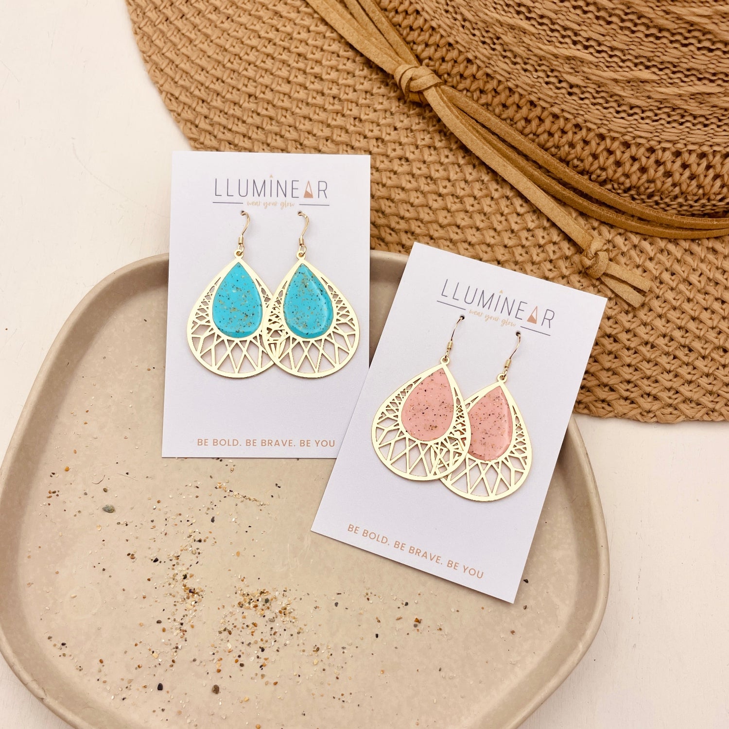 Two pairs of teardrop-shaped earrings displayed on white cards. One pair has a turquoise blue center with a gold filigree border, and the other has a soft pink center with the same gold border.