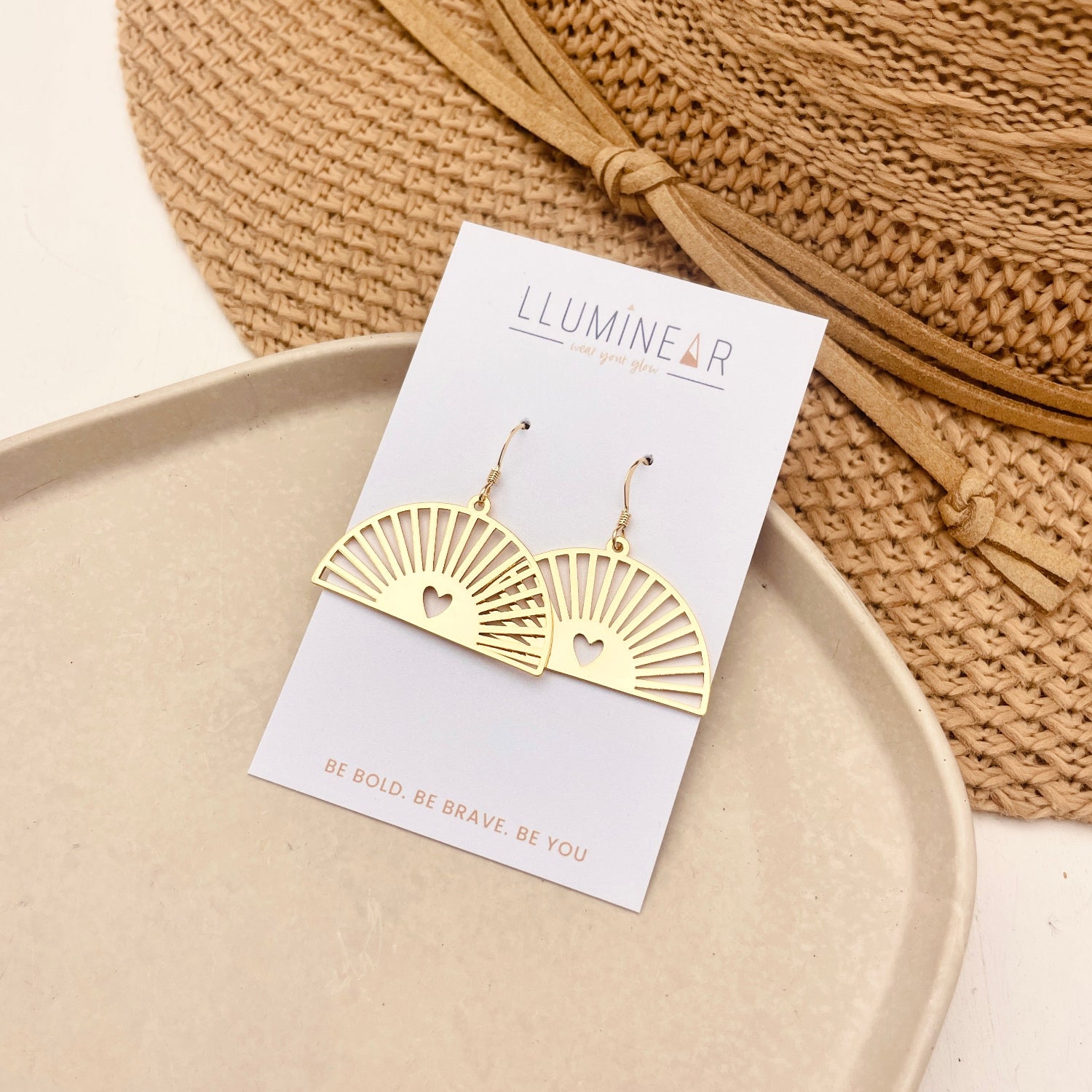 Gold fan-shaped dangle earrings with heart cutouts, displayed on a white card labeled 'LLUMINEAR' and styled on a beige tray with a woven straw hat in the background.