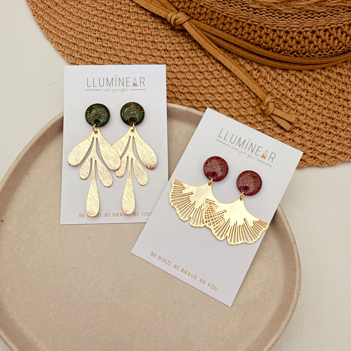 Two pairs of gold-toned statement earrings displayed on white cards. One pair has dark green oval studs with leaf-shaped dangling elements, and the other has deep red oval studs with matching leaf-shaped dangling elements.