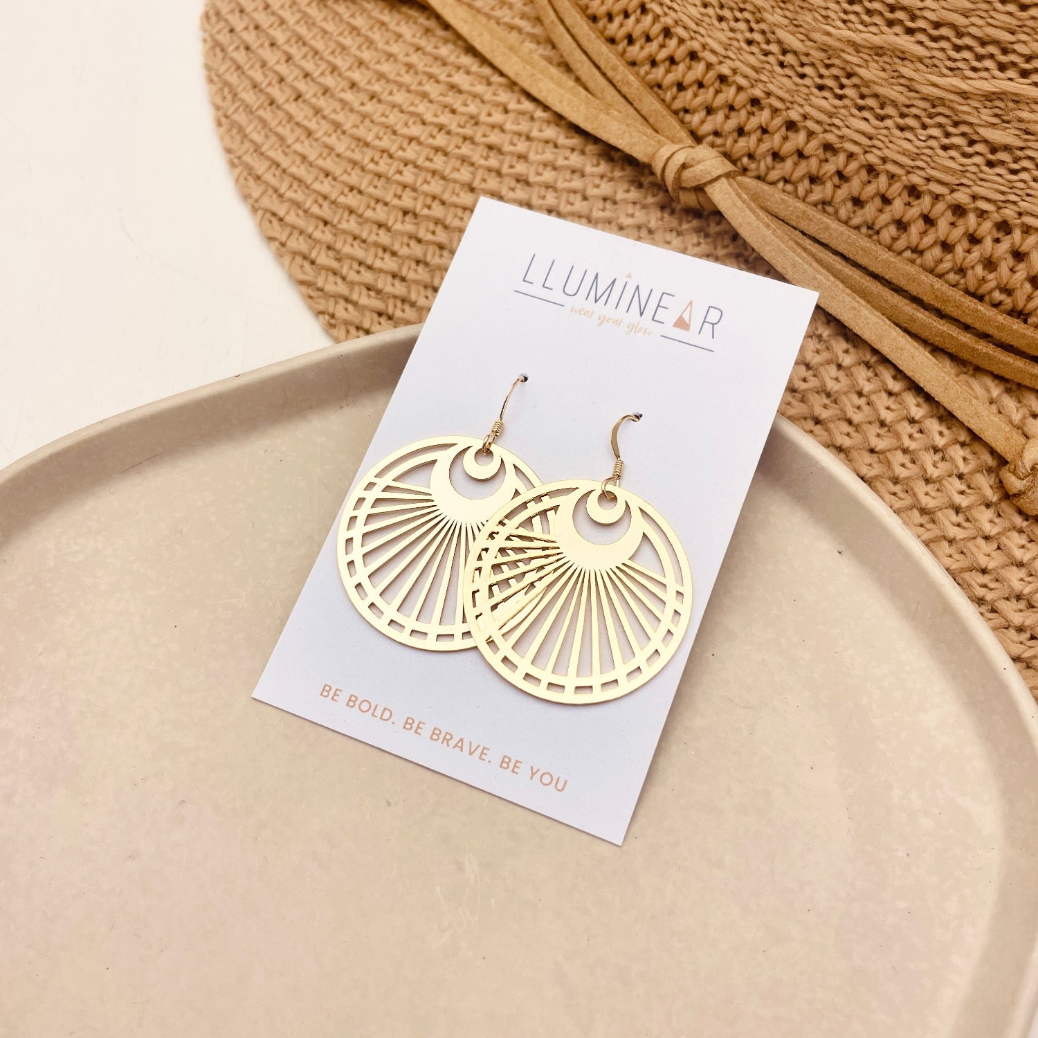A pair of gold-tone dangle earrings displayed on a white card labeled 'ILLUMINAR', placed on a beige tray with a woven straw hat in the background.