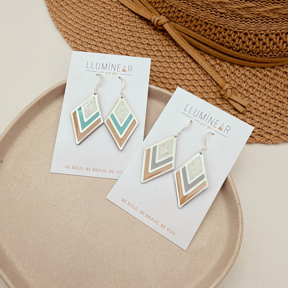 A pair of geometric dangle earrings displayed on white cards, placed on a beige tray with a woven straw hat in the background. The earrings feature chevron patterns in cream, sage green, and light brown, with silver-toned metal hooks.