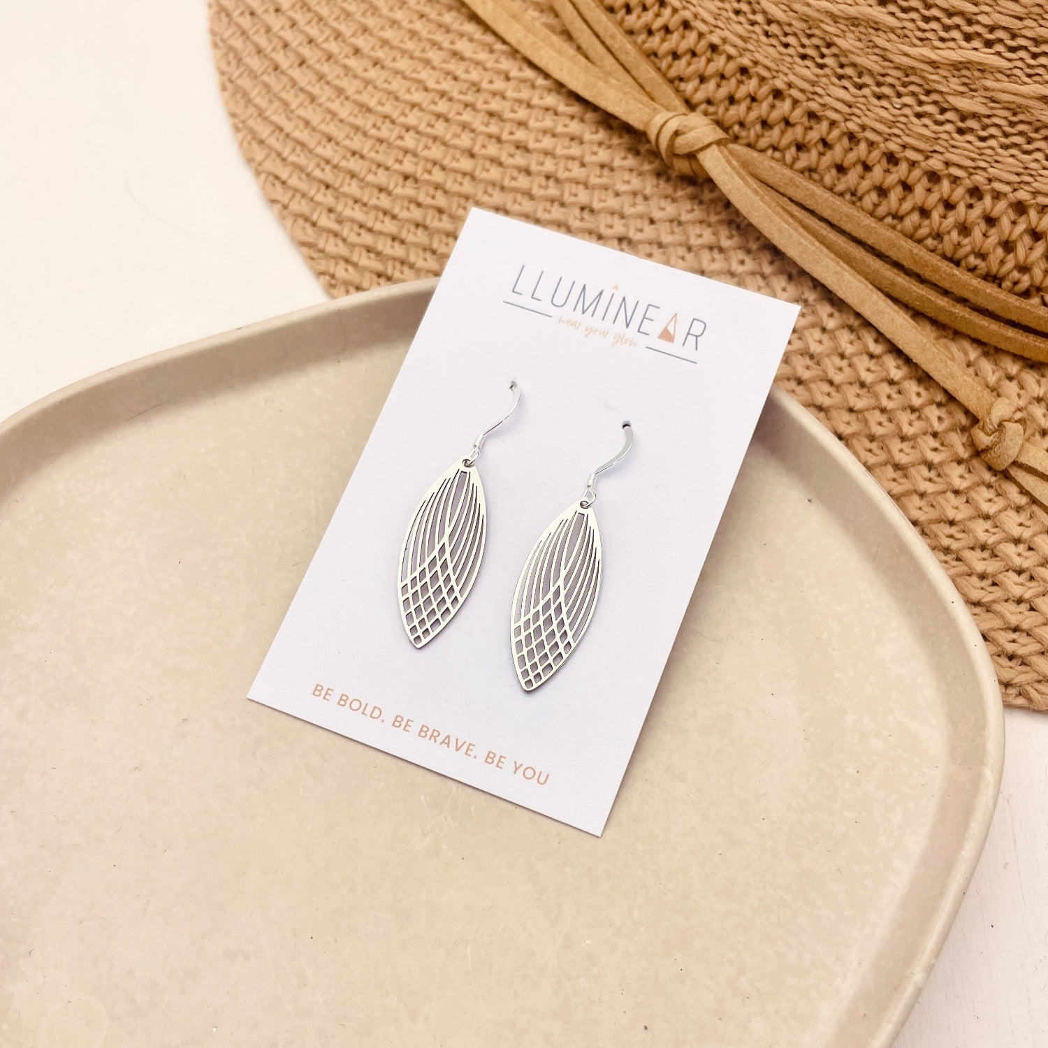 Silver leaf-shaped dangle earrings on a white display card labeled 'LLUMINE&ER', styled on a beige tray with a woven straw hat in the background.