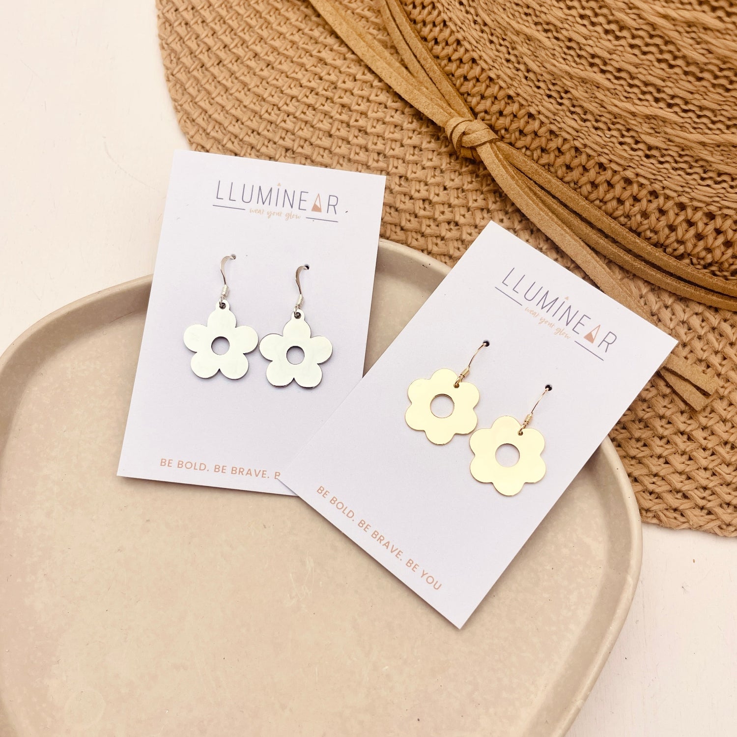 Two pairs of flower-shaped dangle earrings displayed on white cards, one silver-toned and one gold-toned, styled on a beige tray with a woven straw hat in the background.