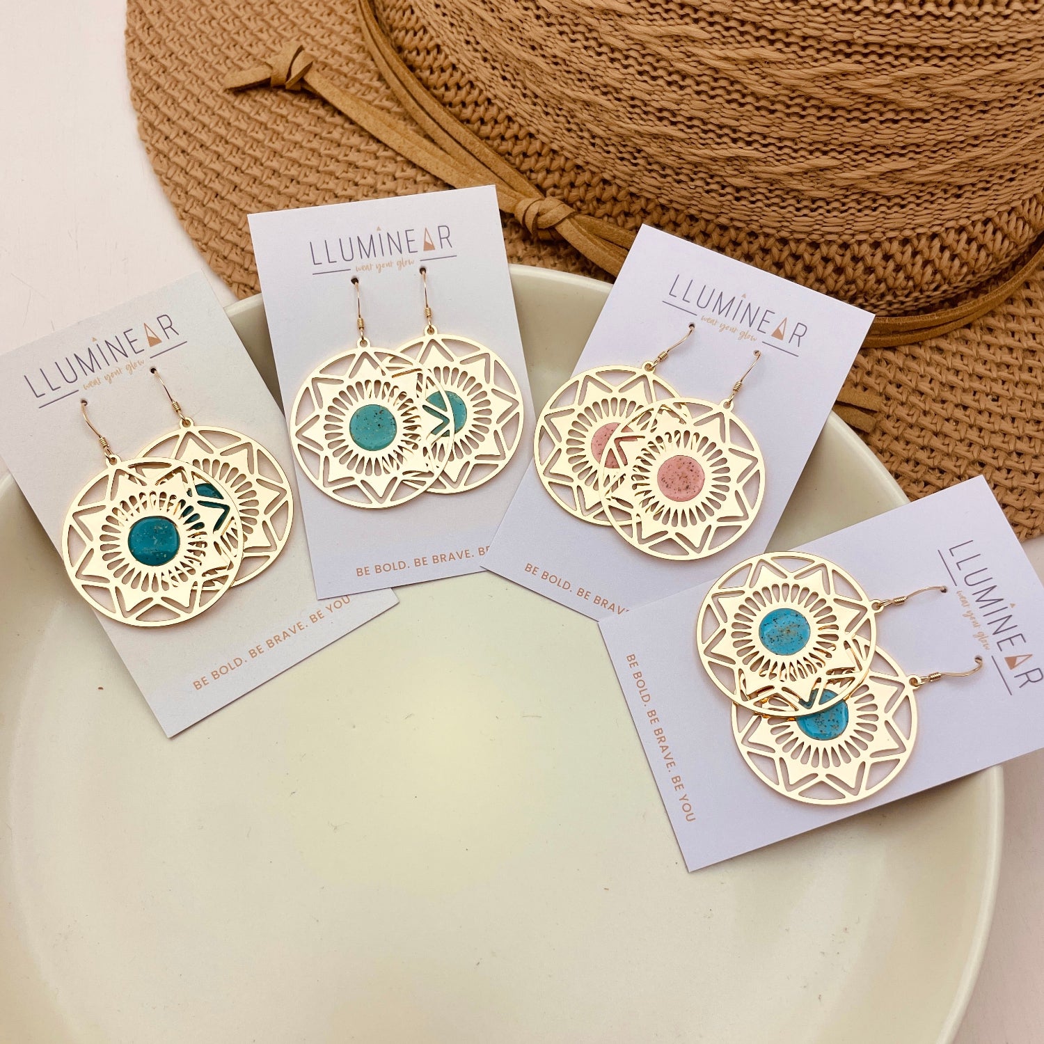 Four pairs of gold-tone geometric dangle earrings displayed on white cards, arranged on a white plate with a woven straw hat in the background. Two pairs feature teal blue stones and two feature peach-pink stones.