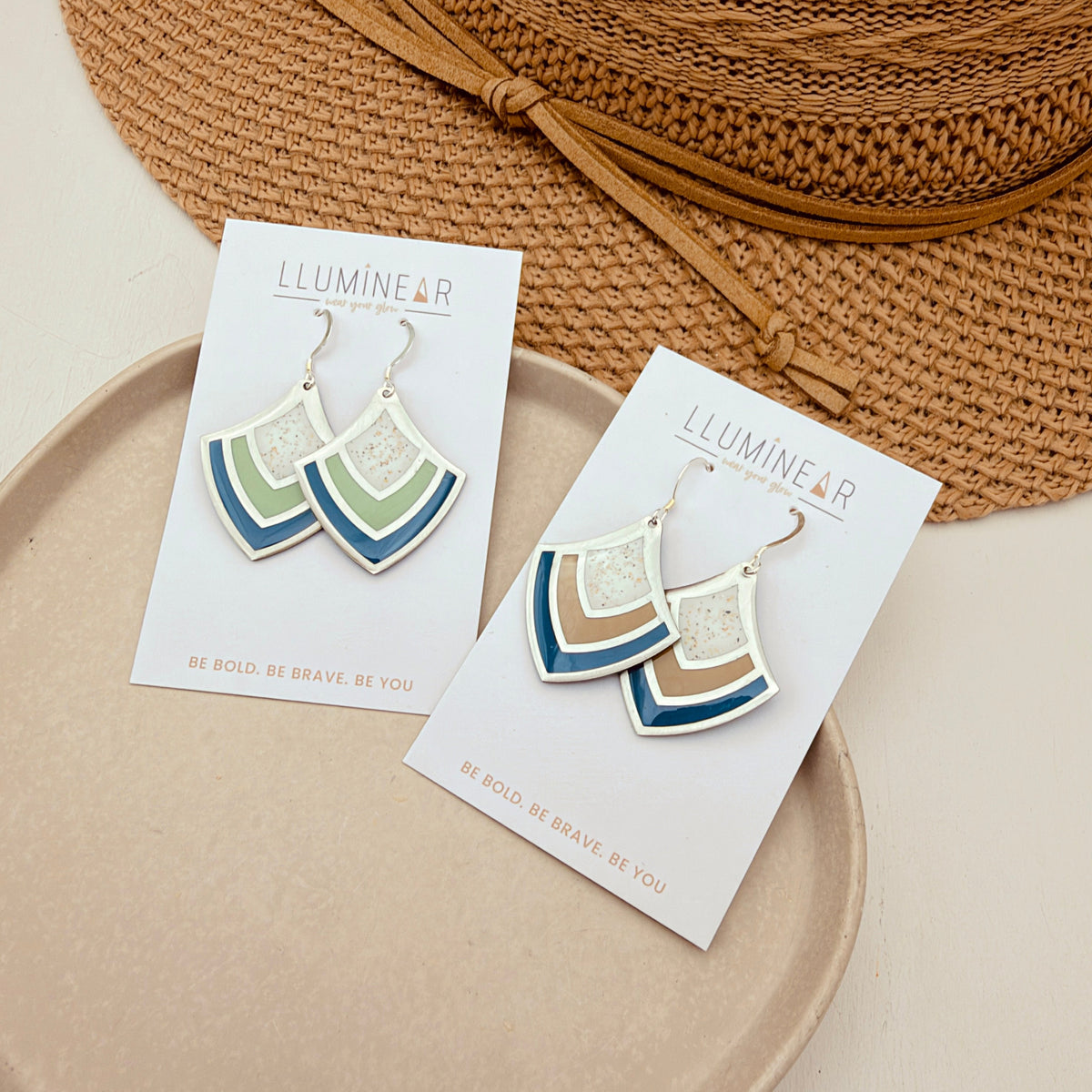 A pair of geometric dangle earrings with white, teal, and tan layers on silver hooks, displayed on white brand cards on a beige tray with a straw hat in the background.