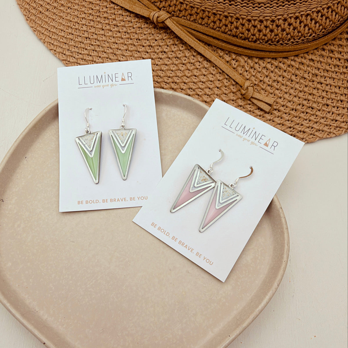 Two pairs of geometric drop earrings displayed on white cards, placed on a beige tray with a woven straw hat behind them. The earrings have chevron designs in mint green, blush pink, and white, with silver-tone hooks.