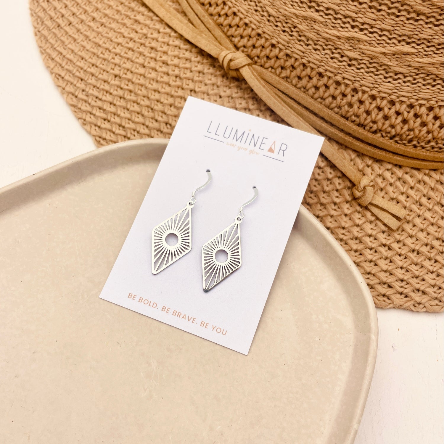 Silver geometric dangle earrings on a white display card, styled on a beige tray with a woven straw hat in the background.