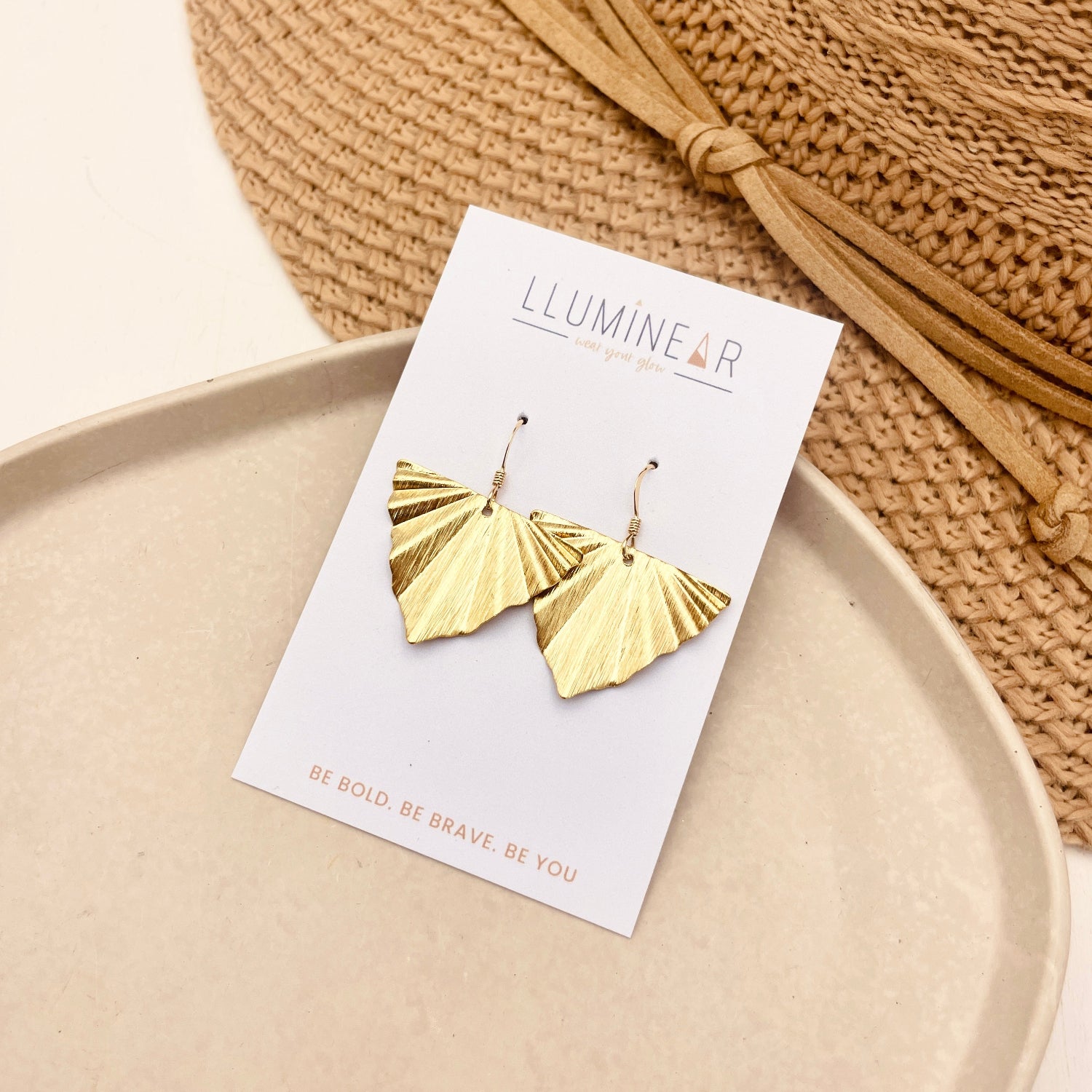 Gold fan-shaped dangle earrings on a white display card, styled on a beige tray with a woven straw hat in the background. The card is branded 'LLUMINE PARIS'.