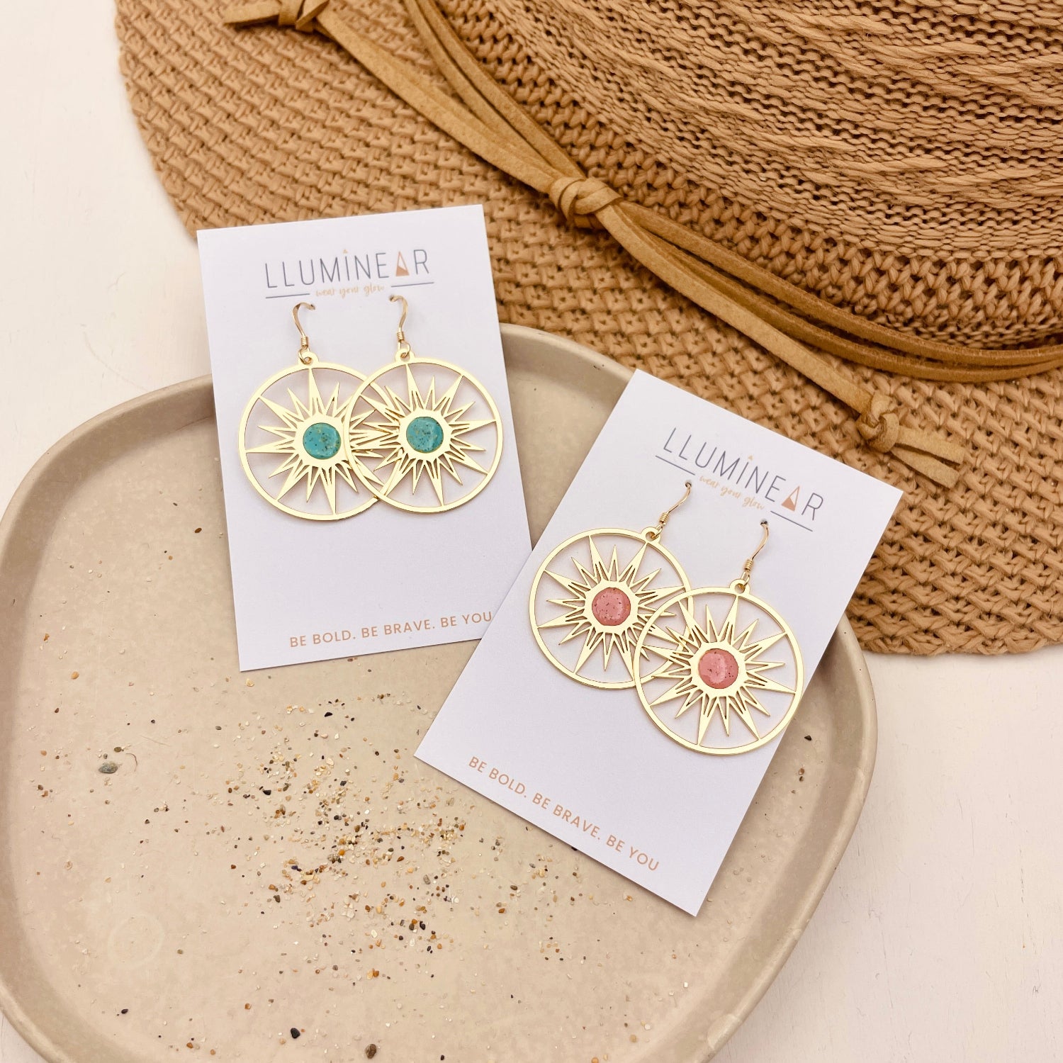 Two pairs of gold-tone sunburst hoop earrings displayed on white cards. One pair has a green stone, and the other has a pink stone, styled on a beige tray with a straw hat.